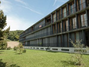 Photo of the Spittelhof Estate apartment building in Biel-Benken, Basel, Switzerland. The building has a long, straight front and is surrounded by green grass and woods.
